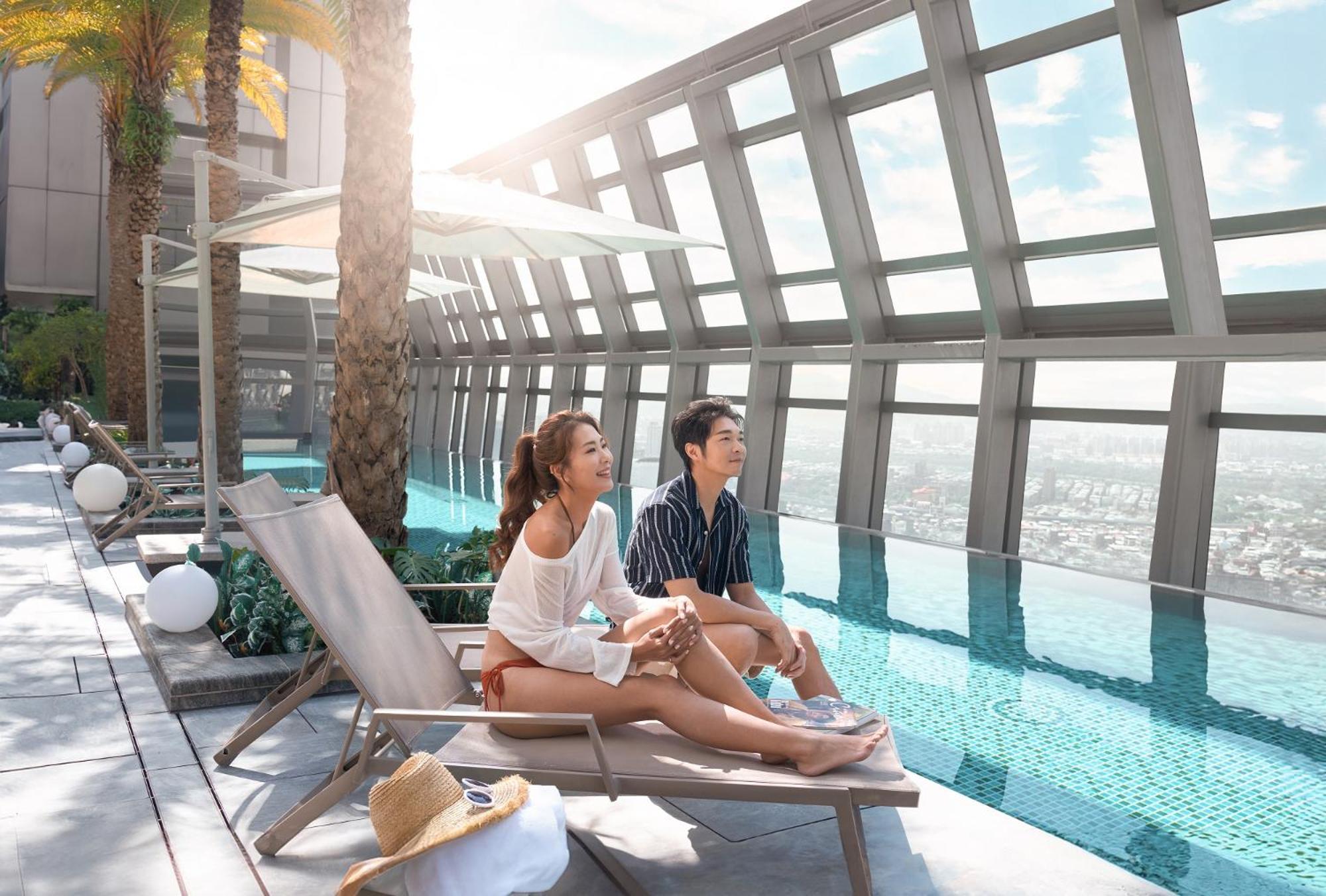 Caesar Park Hotel Banqiao Taipei Exterior photo The photo shows a couple lounging by an infinity pool on a rooftop. They are sitting on deck chairs, enjoying the view, with palm trees in the background. The atmosphere is bright and sunny, and the pool appears to extend out over the edge, creating 