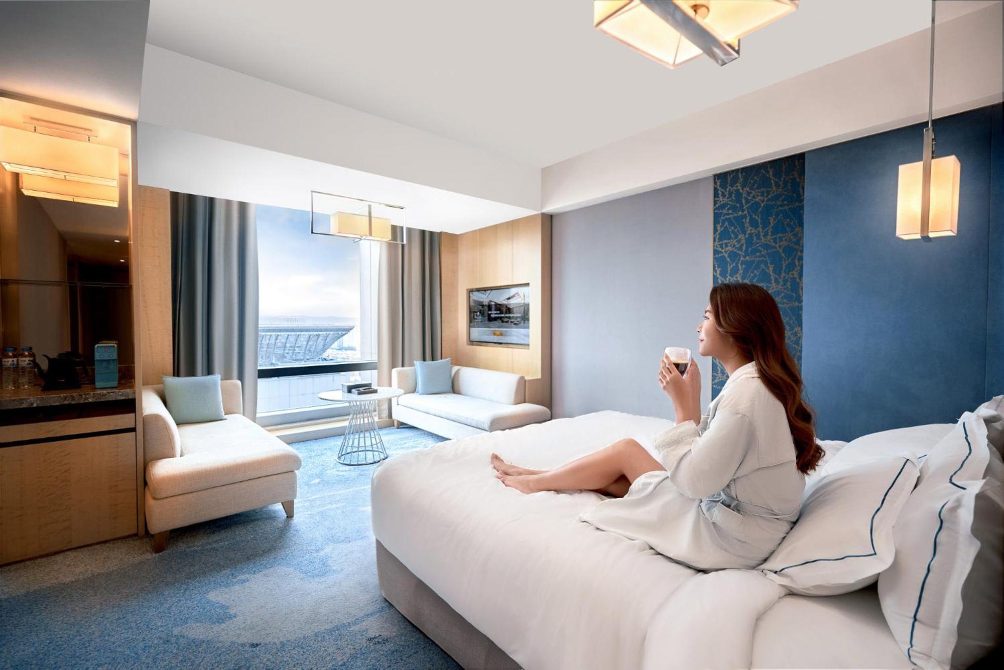 Caesar Park Hotel Banqiao Taipei Exterior photo The photo depicts a modern hotel room featuring a large bed with a light-colored blanket and pillows. A woman in a white robe is sitting on the bed, holding a cup, and appears to be enjoying the serene environment. The room has large windows that off