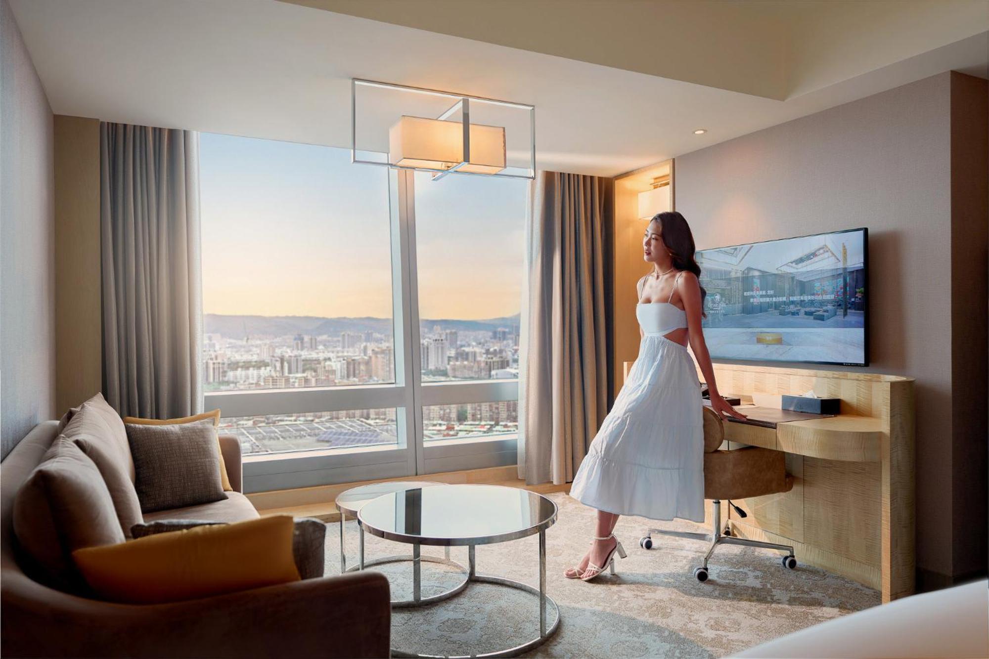 Caesar Park Hotel Banqiao Taipei Exterior photo The photo showcases a modern hotel room with a panoramic view of the city. A woman in a white dress stands next to a stylish table, gazing out of a large window. The room features contemporary furniture, including a sofa, a glass coffee table, and a 