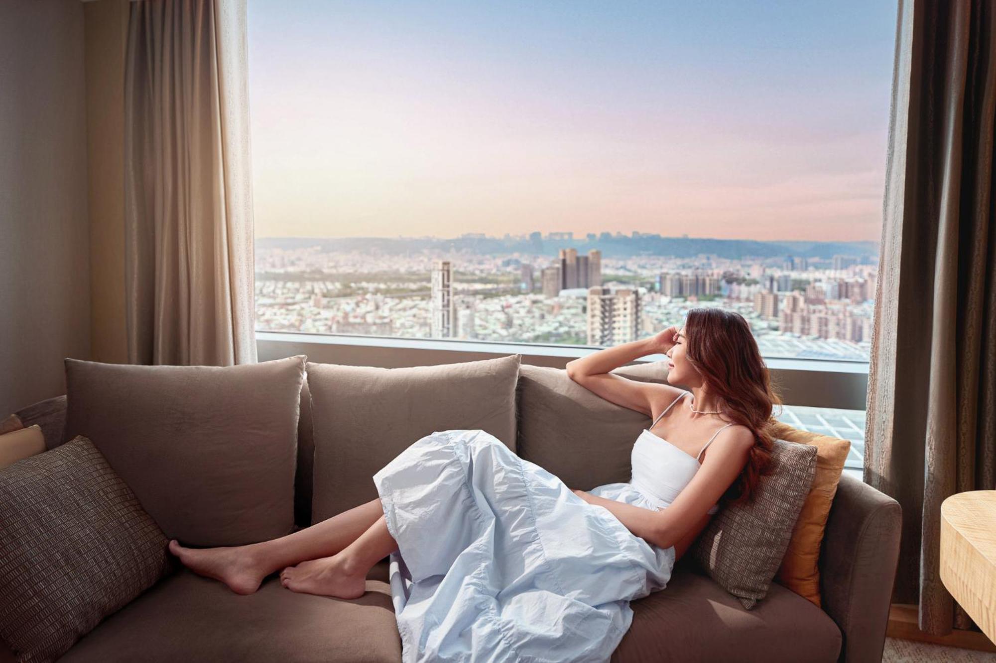 Caesar Park Hotel Banqiao Taipei Exterior photo The image shows a woman sitting on a couch in a bright room, gazing out of a large window that offers a view of a cityscape. She is wearing a white dress and appears to be in a relaxed pose, resting her arm on her head. The window shows a pastel sky,